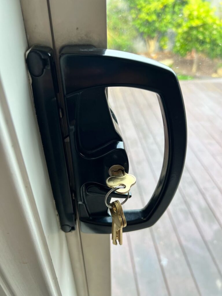 Close-up of a black sliding door lock with keys inserted, mounted on a white door frame with an outdoor view of a wooden deck and greenery.
